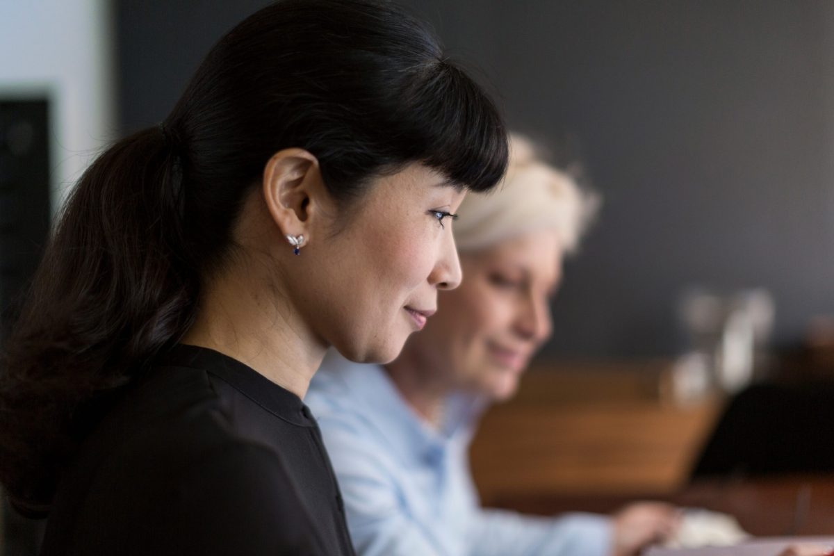 Side view of content Japanese businesswoman sitting at table with diverse female colleague working on project in team